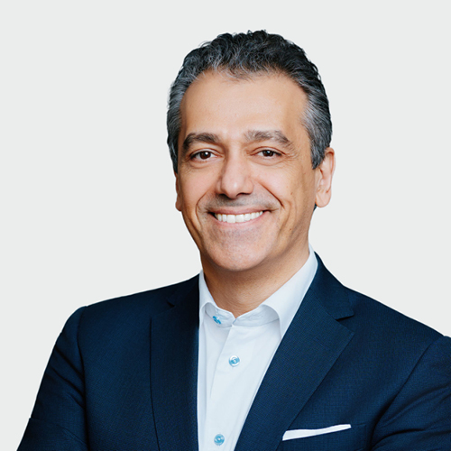 Portrait of a smiling man in a navy suit and white shirt against a light neutral background.