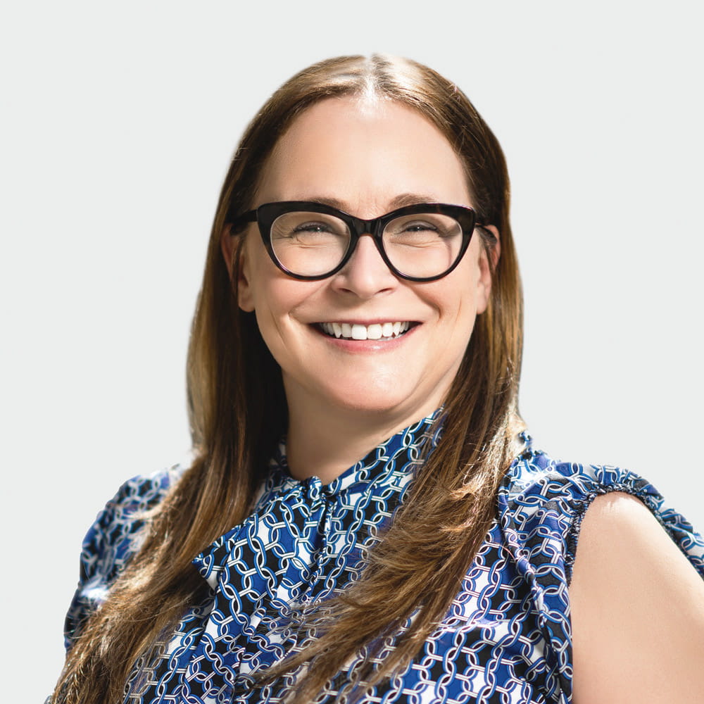 Smiling woman with long brown hair and black glasses, wearing a blue patterned blouse.