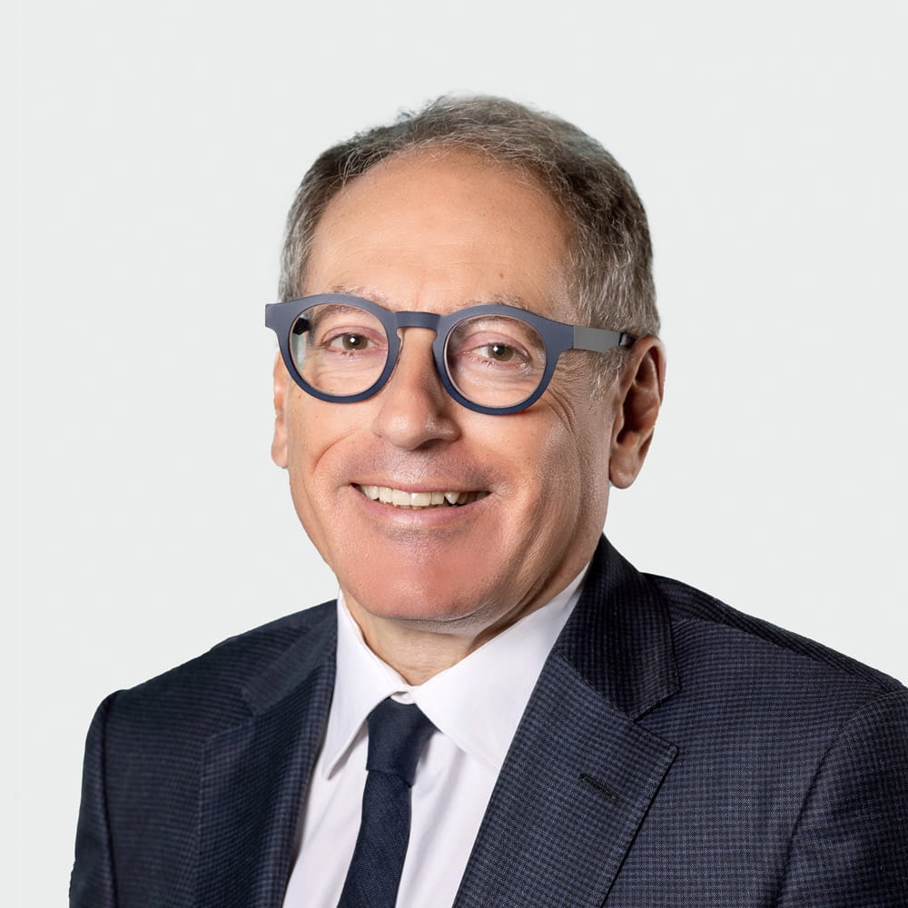 Professional headshot of a smiling man in a dark suit and white shirt, wearing blue round glasses against a light background.