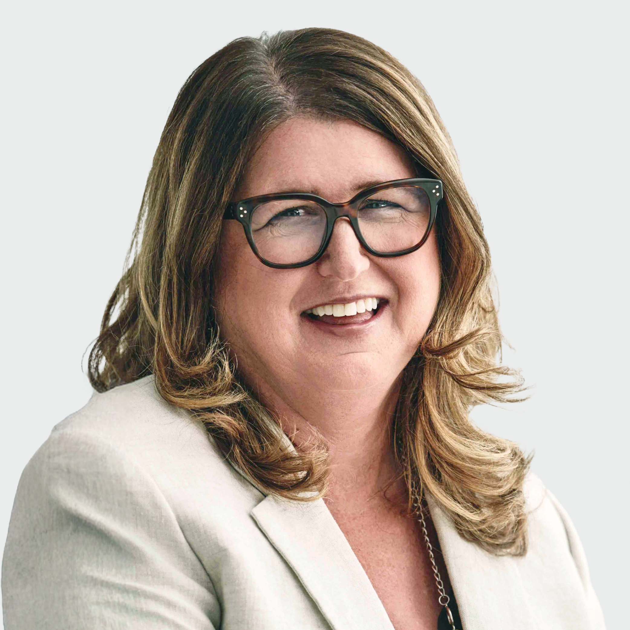 Smiling woman with wavy light-brown hair wearing black glasses and a light blazer, close-up headshot on neutral background.