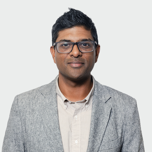 Professional headshot of a man with short dark hair and glasses, wearing a gray blazer and light checkered shirt against a light neutral background.