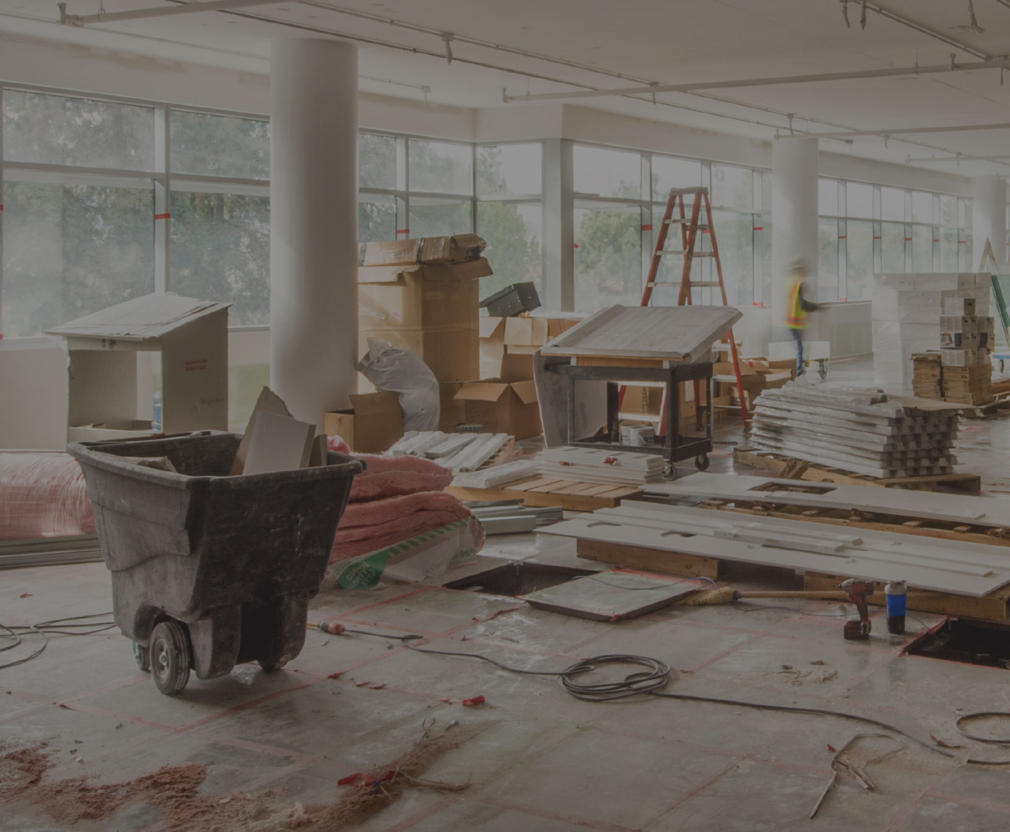 Indoor construction site with a wheelbarrow, scattered boxes and materials, and a worker in a safety vest in the background.