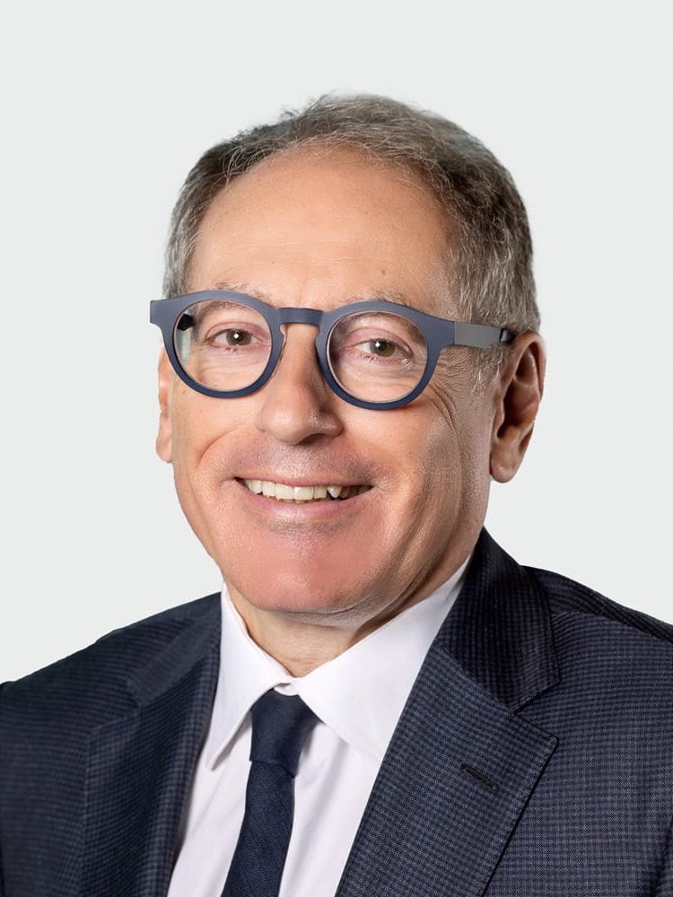 Professional headshot of a smiling man in a dark suit and white shirt, wearing blue round glasses against a light background.