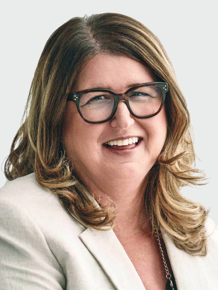 Smiling woman with wavy light-brown hair wearing black glasses and a light blazer, close-up headshot on neutral background.