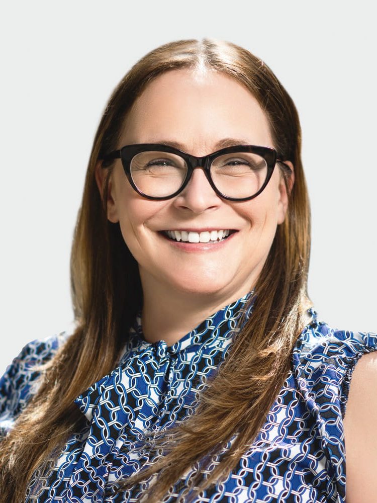 Smiling woman with long brown hair and black glasses, wearing a blue patterned blouse.