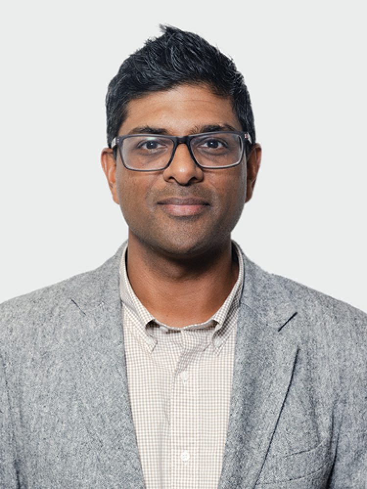 Professional headshot of a man with short dark hair and glasses, wearing a gray blazer and light checkered shirt against a light neutral background.